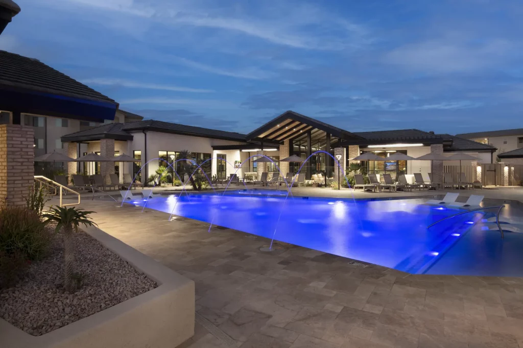 Resort pool at Encantada Gladden Farms illuminated at dusk, featuring blue underwater lighting, arching water jets, lounge chairs with umbrellas, and a covered clubhouse in the background.